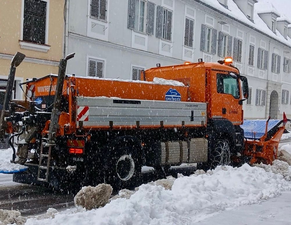 Ein orangefarbener Schneepflug mit Springer-Logo räumt Schnee von einer Straße. Das Fahrzeug ist am Straßenrand geparkt, der Boden ist schneebedeckt. Dahinter steht ein Gebäude mit vielen Fenstern.