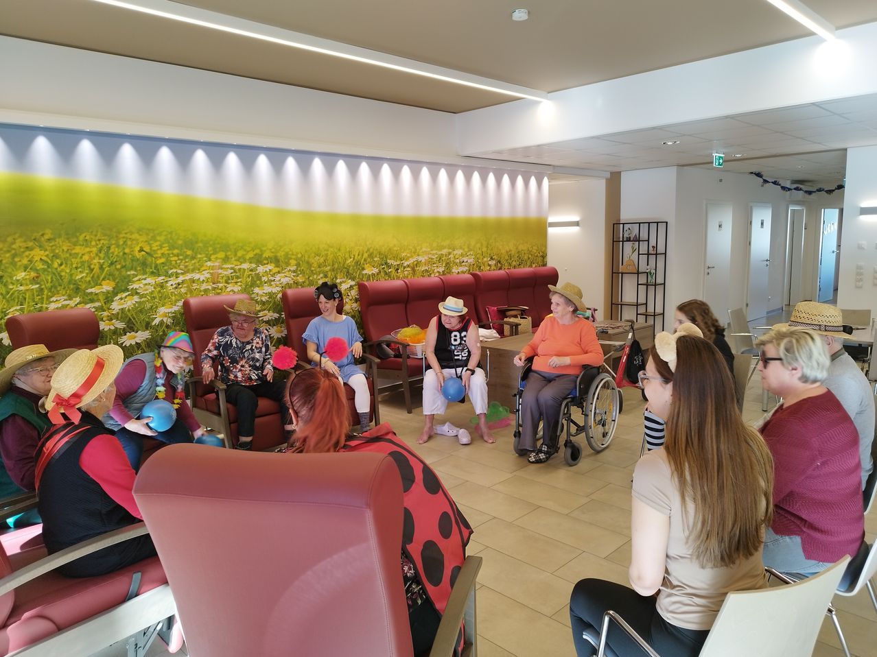Eine Gruppe älterer Frauen in Hüten sitzt in roten Stühlen um einen Raum mit einem Blumenfeld-Wandbild. Eine Frau ist im Rollstuhl und alle halten bunte Bälle.