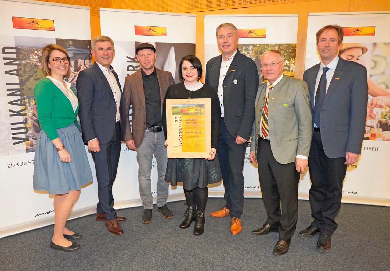 Eine Gruppe von Menschen steht lächelnd vor einer Kulisse. Eine Frau in der Mitte hält ein gelbes Zertifikat. Sie sind formell gekleidet. Dahinter befinden sich Banner mit Logos und Text.