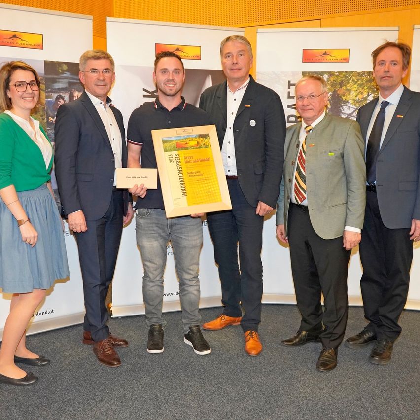 Eine Gruppe von Personen steht im Innenraum und lächelt. Ein Mann in der Mitte hält ein Zertifikat. Der Mann auf der linken Seite trägt ein grünes Hemd und der Mann auf der rechten Seite trägt eine Krawatte. Hinter ihnen befinden sich Banner mit Logos und Text.