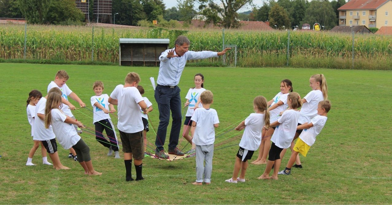 Ein Mann steht auf einem Brett und balanciert, während er Seile hält, die mit Kindern in weißen Shirts verbunden sind, auf einem Rasenfeld mit Zaun und Bäumen im Hintergrund.