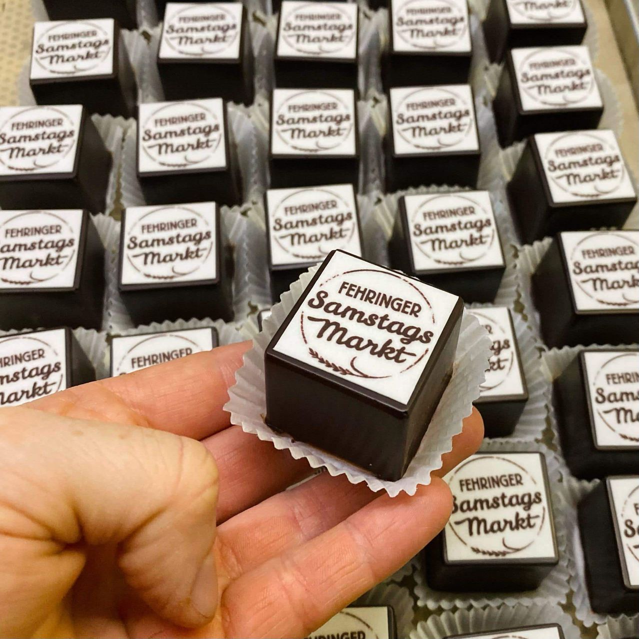 Eine Hand hält eine Schokoladentafel mit der Aufschrift 'Fehringer Samstags Markt'. Dahinter befinden sich zahlreiche ähnliche Schokoladen in Verpackungen, jede mit der gleichen Bezeichnung, in einer Reihe angeordnet.