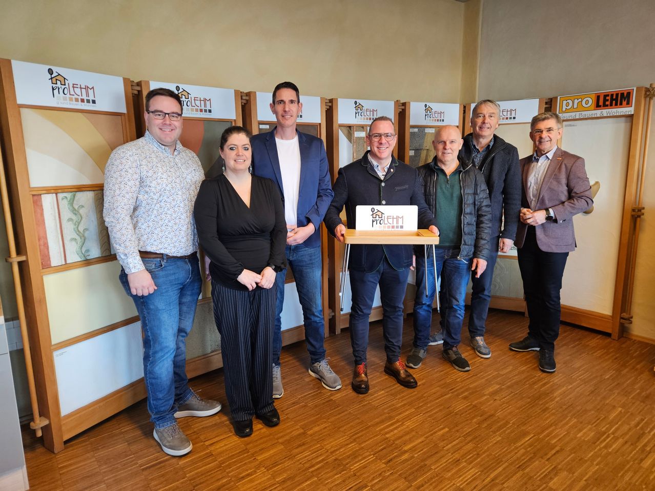 Eine Gruppe von Menschen steht lächelnd in einem Raum zusammen. Ein Mann hält ein Schild mit einem Logo. Hinter ihnen haben die Wände Logos und Designs. Der Boden ist aus Holz.