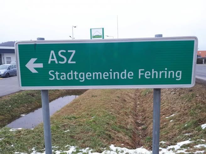 Ein grünes Straßenschild mit weißem Text zeigt nach links zu ASZ Stadtgemeinde Fehring. Das Schild ist an einem Metallpfosten montiert. Dahinter befindet sich ein kleiner Bach und ein geparktes Auto an der Seite.