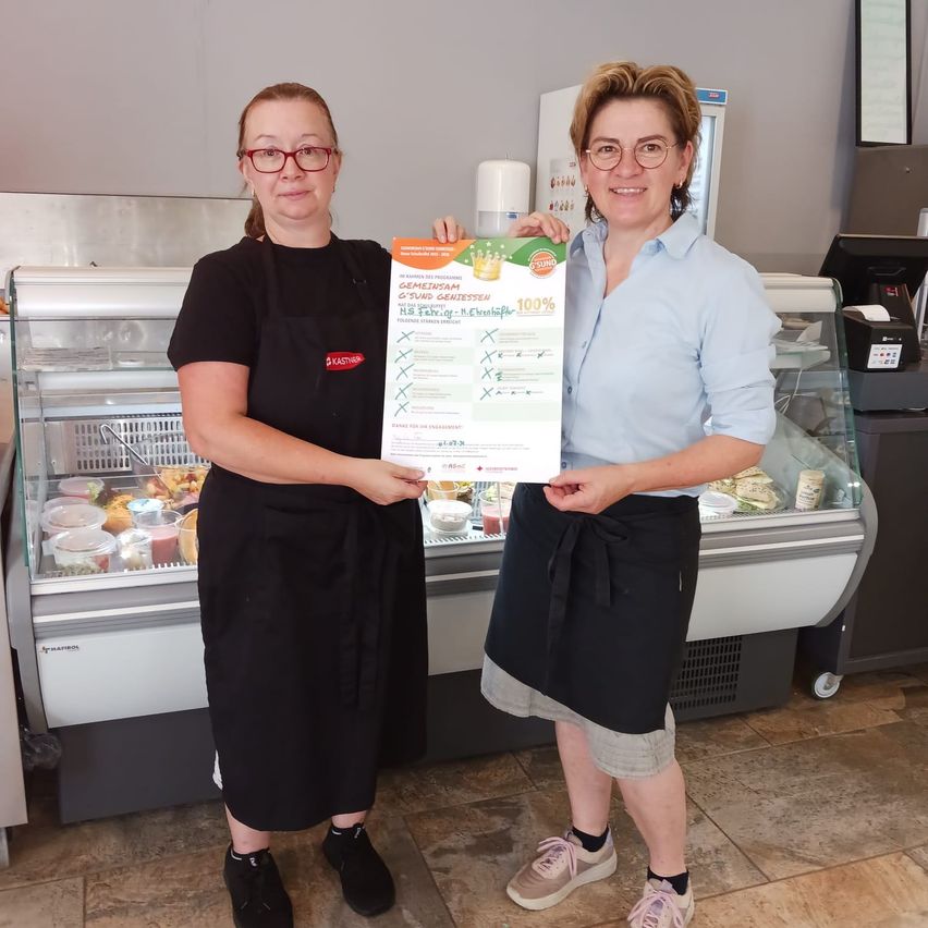 Zwei Frauen stehen vor einer Vitrine in einem Laden, eine hält ein Plakat mit der Aufschrift '100% frisch'.
