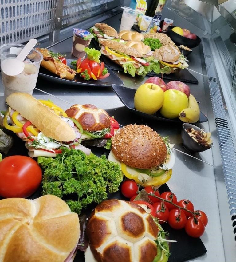 Eine Auswahl an Sandwiches, einschließlich eines Kreuzbrötchens, wird auf einem Tresen mit frischem Gemüse und Obst präsentiert. Es gibt auch Joghurts und ein Getränk in einem Becher.