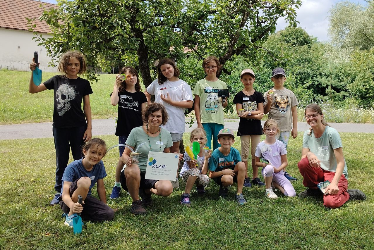 Eine Gruppe von Kindern und Erwachsenen posiert für ein Foto in einem Park mit einem Baum im Hintergrund. Sie halten verschiedene Gegenstände in den Händen und einige tragen Brillen. Eine Frau in der Mitte hält ein Schild mit dem Wort 'KLAR!' darauf.