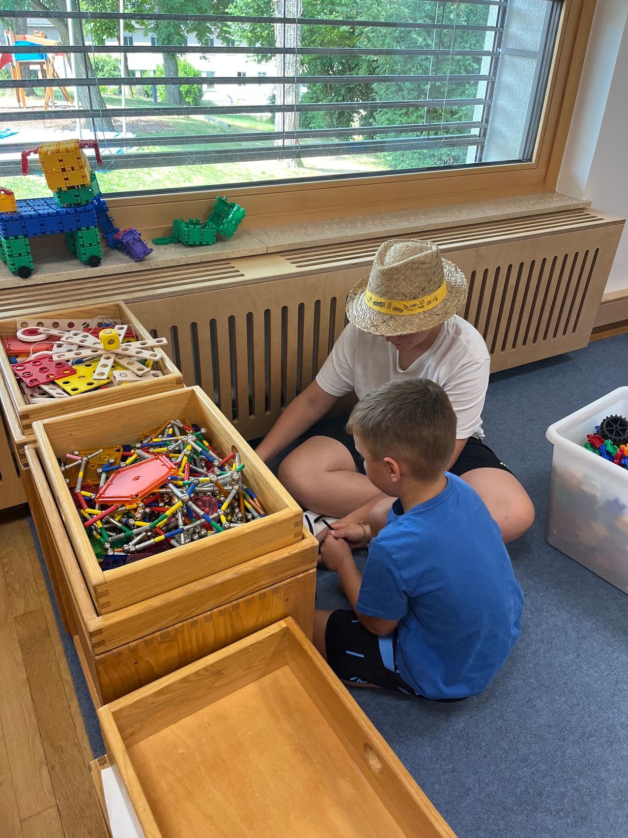 Ein Junge und ein Mann sitzen auf dem Boden und spielen mit bunten Bauklötzen. Der Junge trägt ein blaues T-Shirt und Shorts. Der Mann trägt einen Strohhut und ein weißes T-Shirt. Es gibt Holzkisten voller Bauklötze. Hinter ihnen befindet sich ein Fenster mit Jalousien und einem Heizkörper.