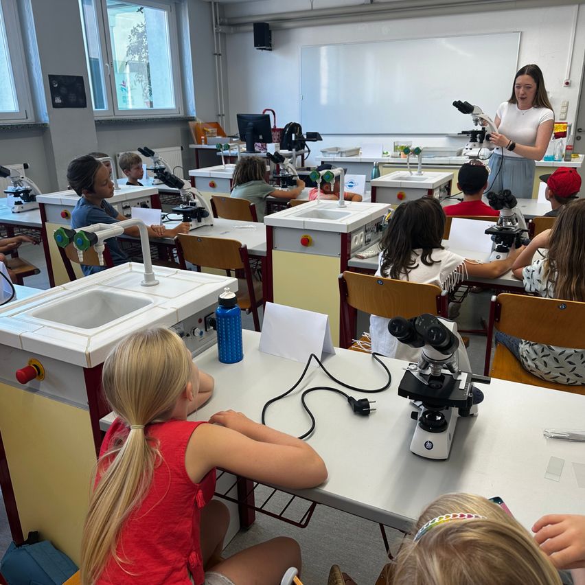 Ein Klassenzimmer mit Schülern, die Mikroskope an ihren Tischen benutzen, eine Lehrerin steht vor einer Tafel.