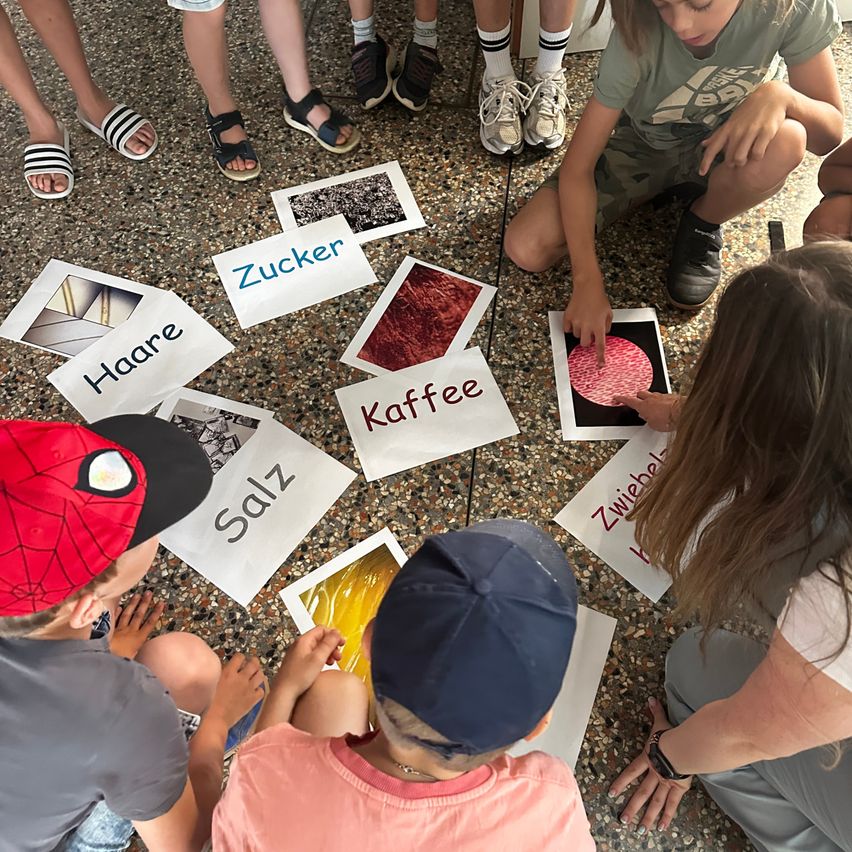 Eine Gruppe von Kindern sitzt im Kreis auf dem Boden, jedes hält ein Stück Papier mit einem darauf geschriebenen Wort. Die Wörter sind 'Zucker', 'Kaffee', 'Salz' und 'Haare'. Die Kinder betrachten ein Bild eines roten Objekts.