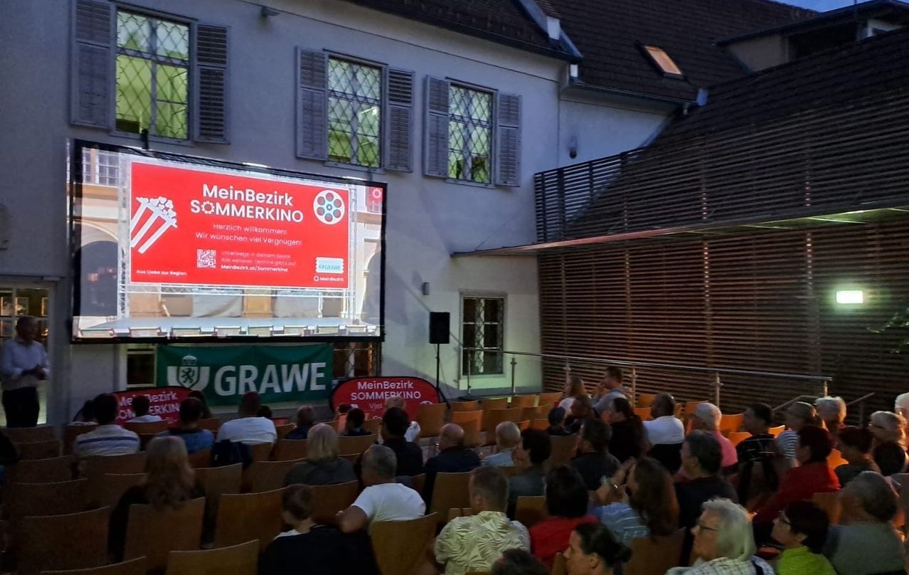 Eine Freiluft-Filmvorführung, 'MeinBezirk Sommerkino', mit einer großen Leinwand und einer Menge Menschen, die auf Stühlen sitzen.