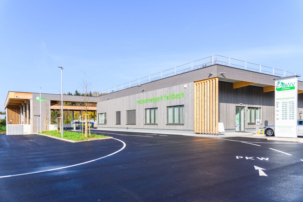 Ein modernes Gebäude mit einem grünen Schild, das 'resourcepark feldbach' besagt. Das Gebäude hat ein flaches Dach mit einer Metallbrüstung, großen Fenstern und einem hölzernen Eingang. Der Parkplatz davor ist leer, mit einer weißen Linie, die die Parkplätze markiert.