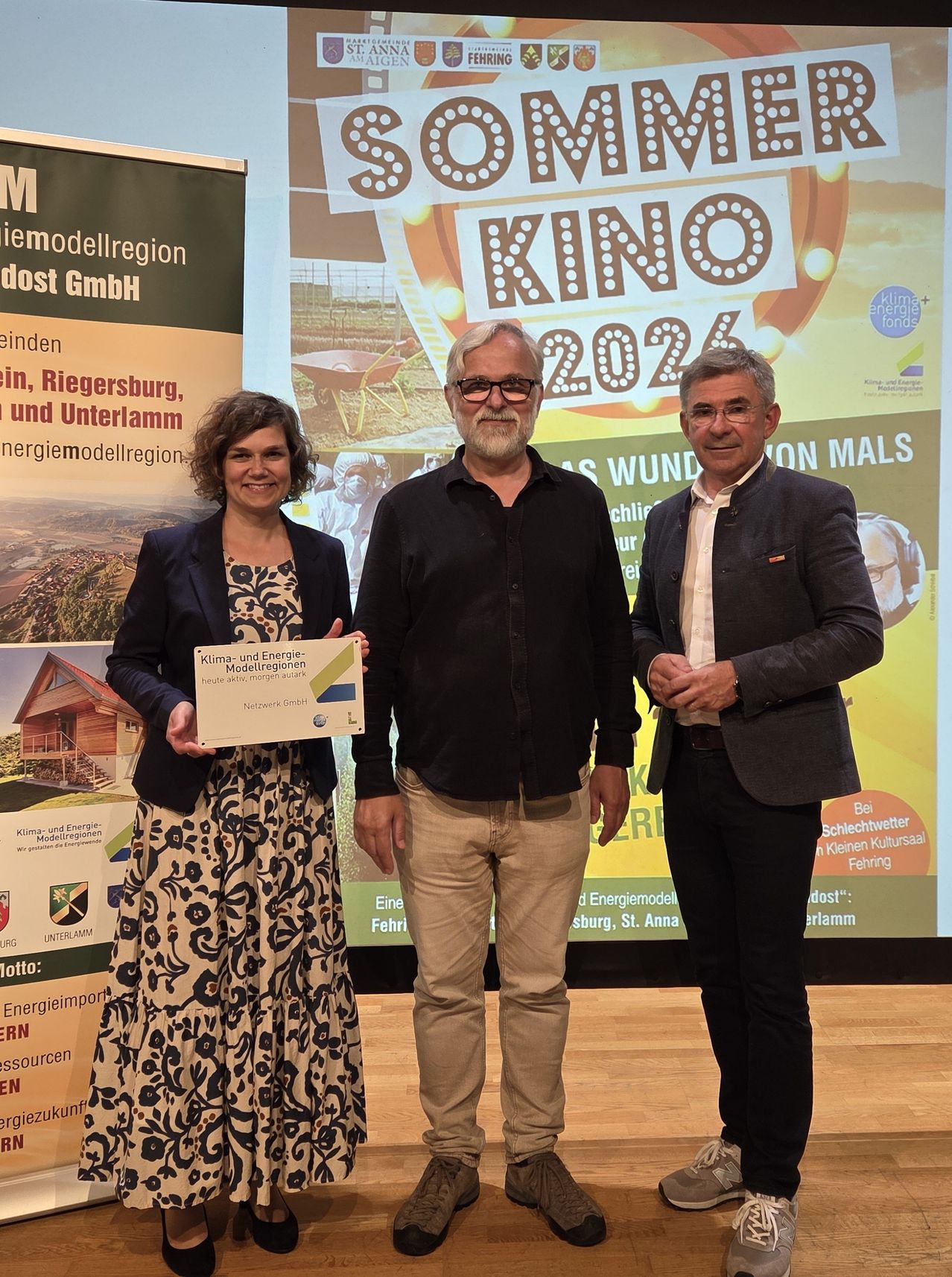 Three individuals stand in front of a large screen with a presentation about 'Klima und Energie-Modellregionen'. The woman holds a certificate, while the two men stand beside her smiling.