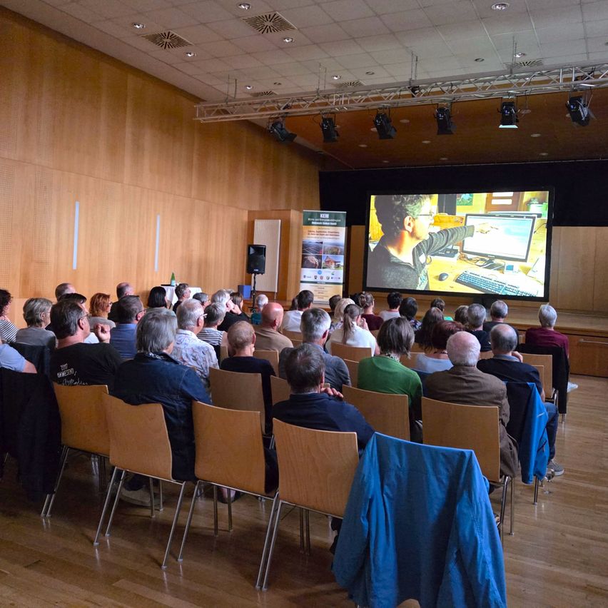 A group of people are seated in chairs in a conference hall, watching a presentation on a large screen. The room has wooden walls and floors, and there is a stage with a computer setup.