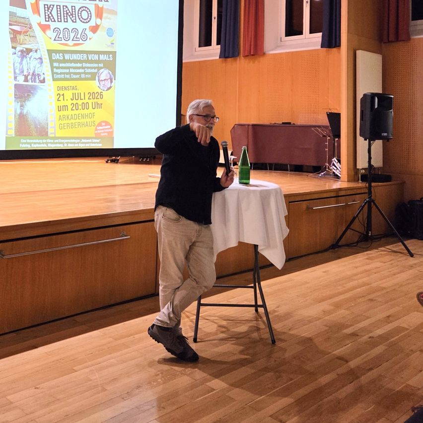 An older man stands at a table with a microphone and a bottle, giving a speech in a room with a large screen showing a movie poster.