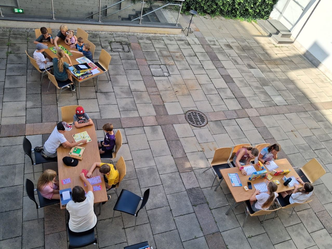 A group of children and adults are playing board games on a brick patio. The patio is outdoors with a staircase and railings visible in the background.