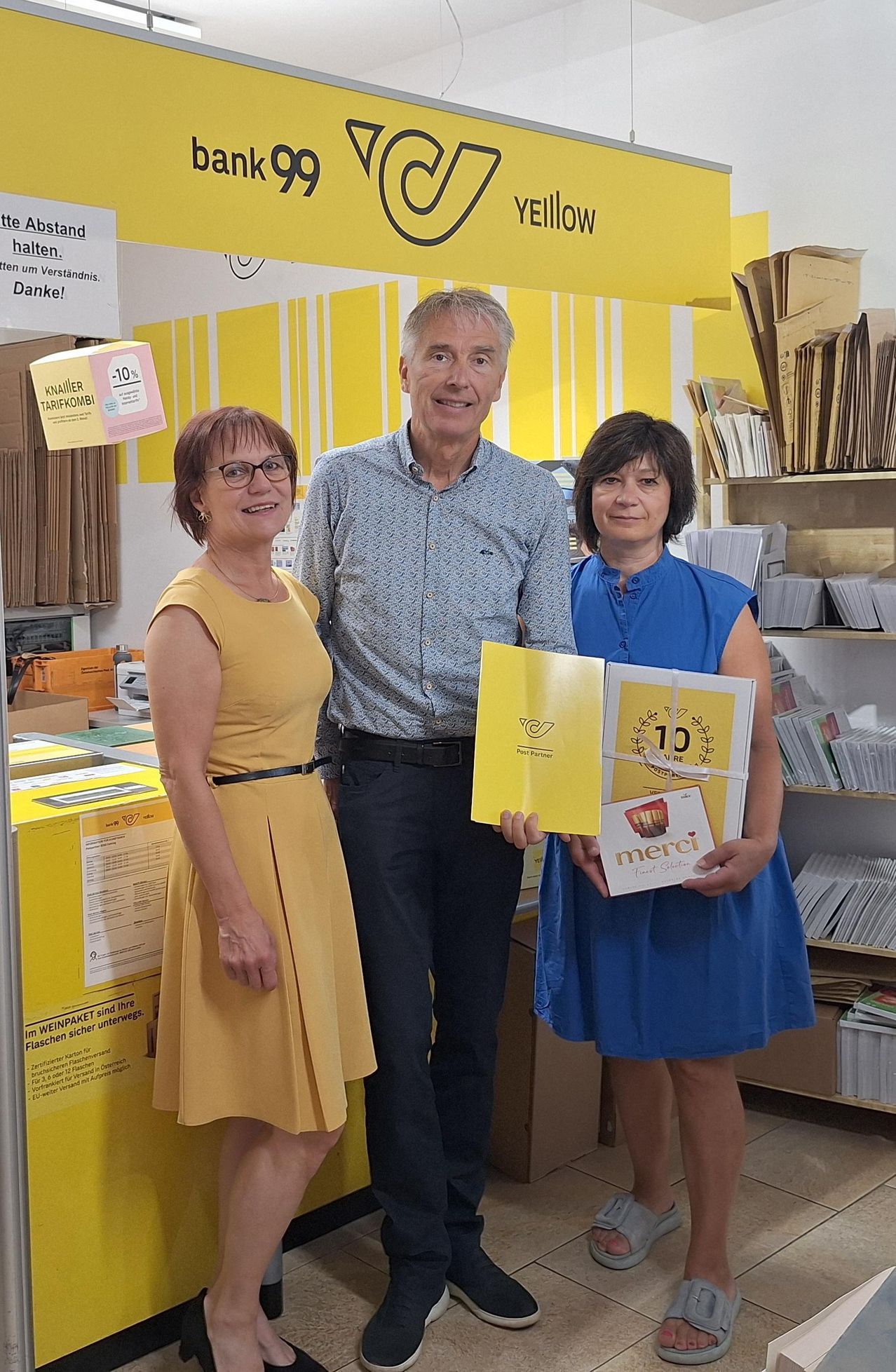 Three people stand together in front of a yellow backdrop with a sign that reads '10%'. The woman on the left wears a yellow dress, the man in the middle a blue shirt, and the woman on the right a blue dress. They hold yellow booklets with the word 'merci' on them. Behind them, shelves filled with books and boxes are visible.