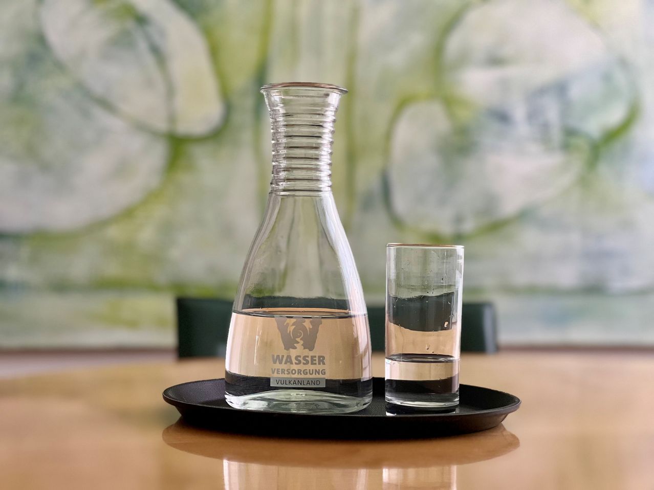 A clear glass water carafe with a label reading 'Wasser Versorgung Vulkanland' and a glass on a black tray on a wooden table.