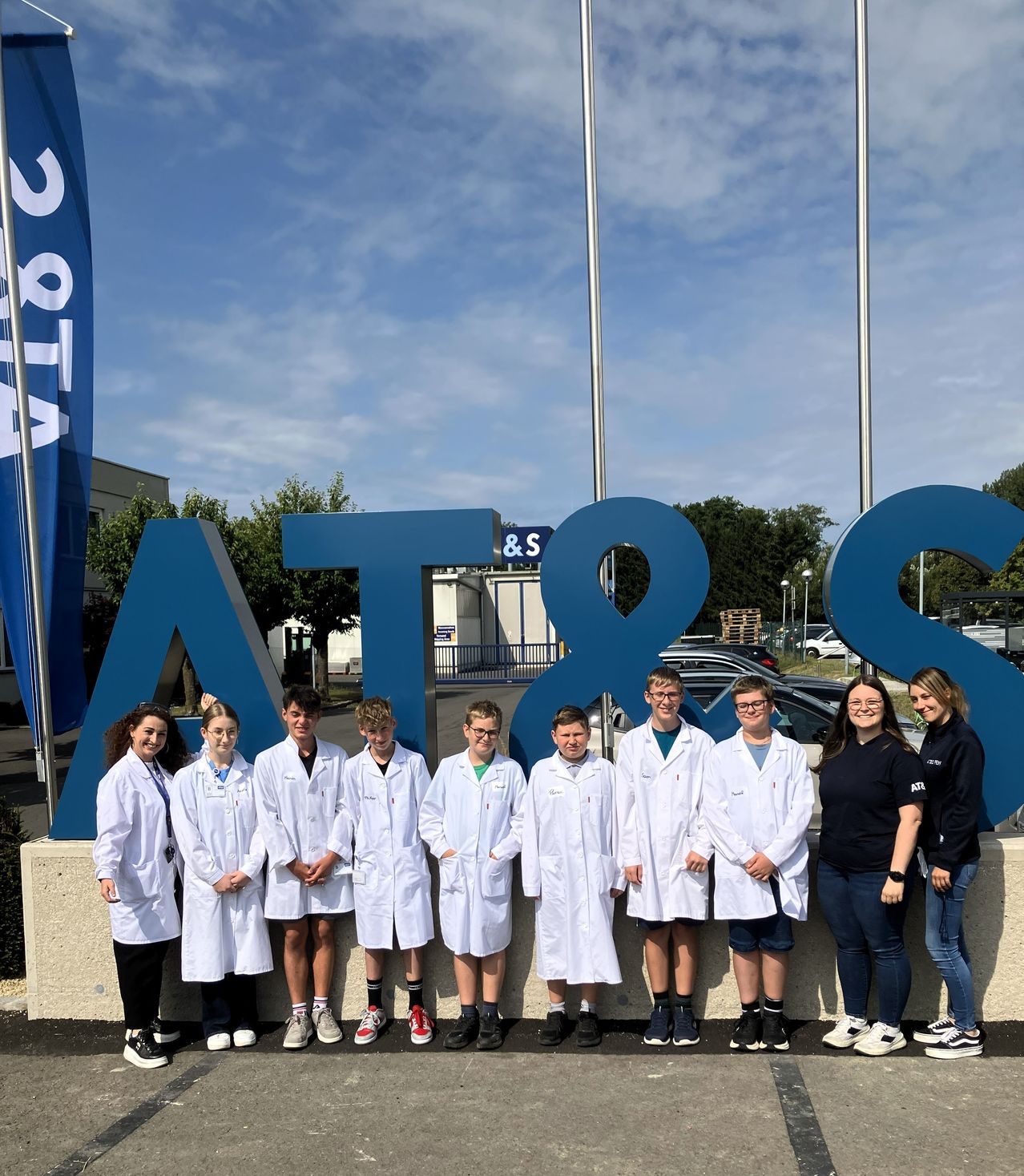A group of young students in white lab coats stand in front of a large blue sign with the letters AT&S.
