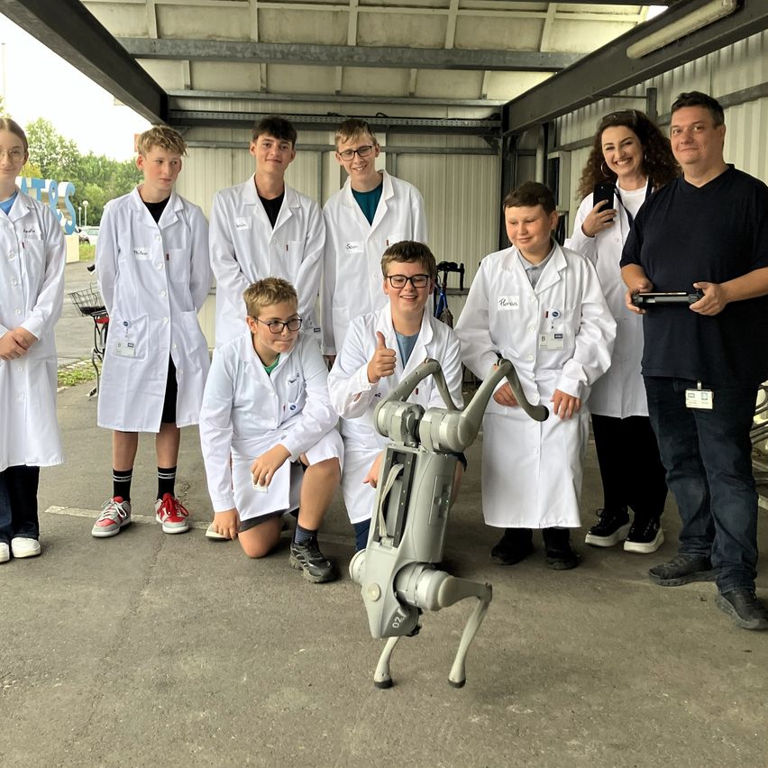 A group of young students in white lab coats are posing with a robot while two adults stand nearby.