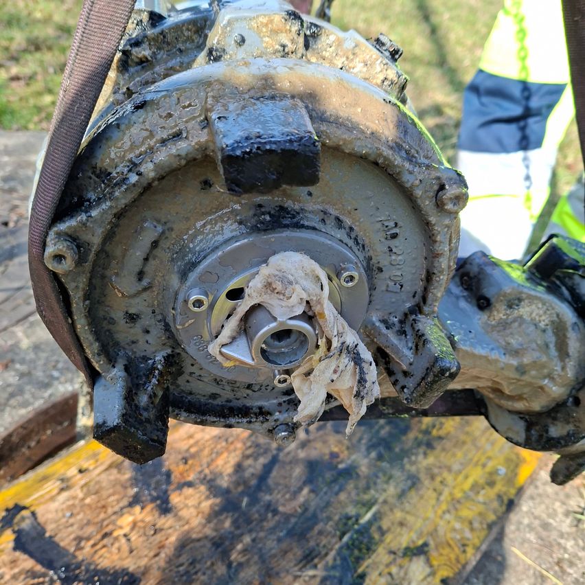 A close-up of a dirty mechanical part with a cloth wrapped around its center, resting on a wooden surface.