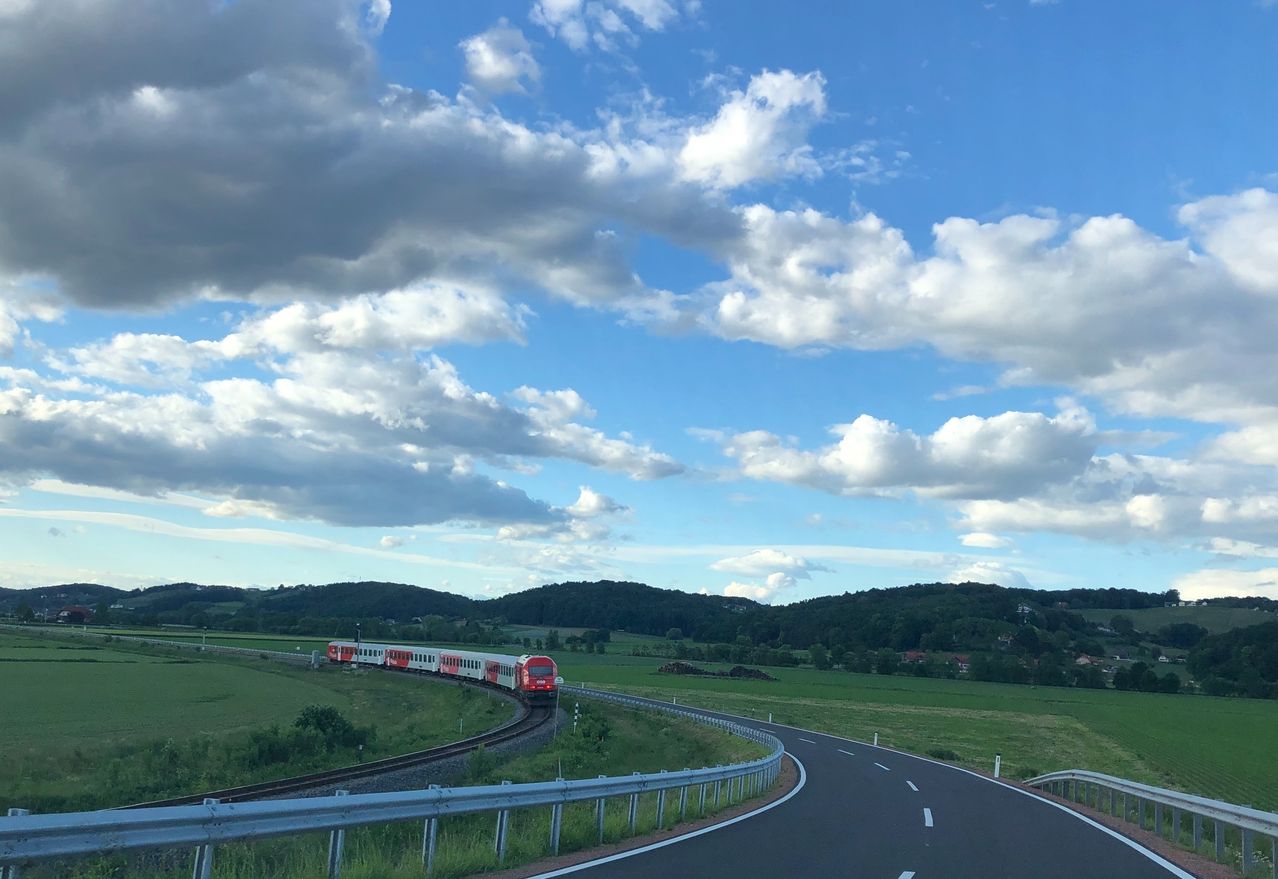 A red and white train travels on a track through a green landscape under a partly cloudy sky.