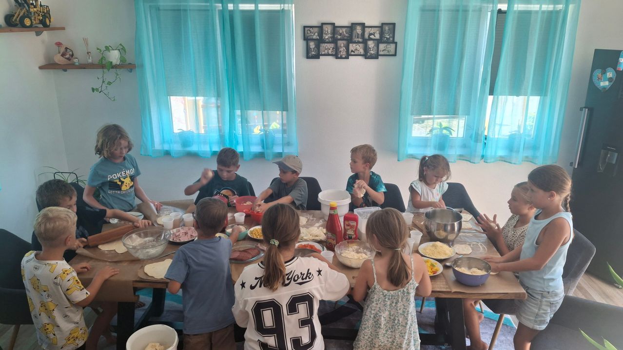 A group of children are gathered around a table, preparing food. They are surrounded by various ingredients and utensils. The table is set with bowls, plates, and cups. The children are wearing casual clothing. In the background, there is a wall with a picture frame and a window with blue curtains.