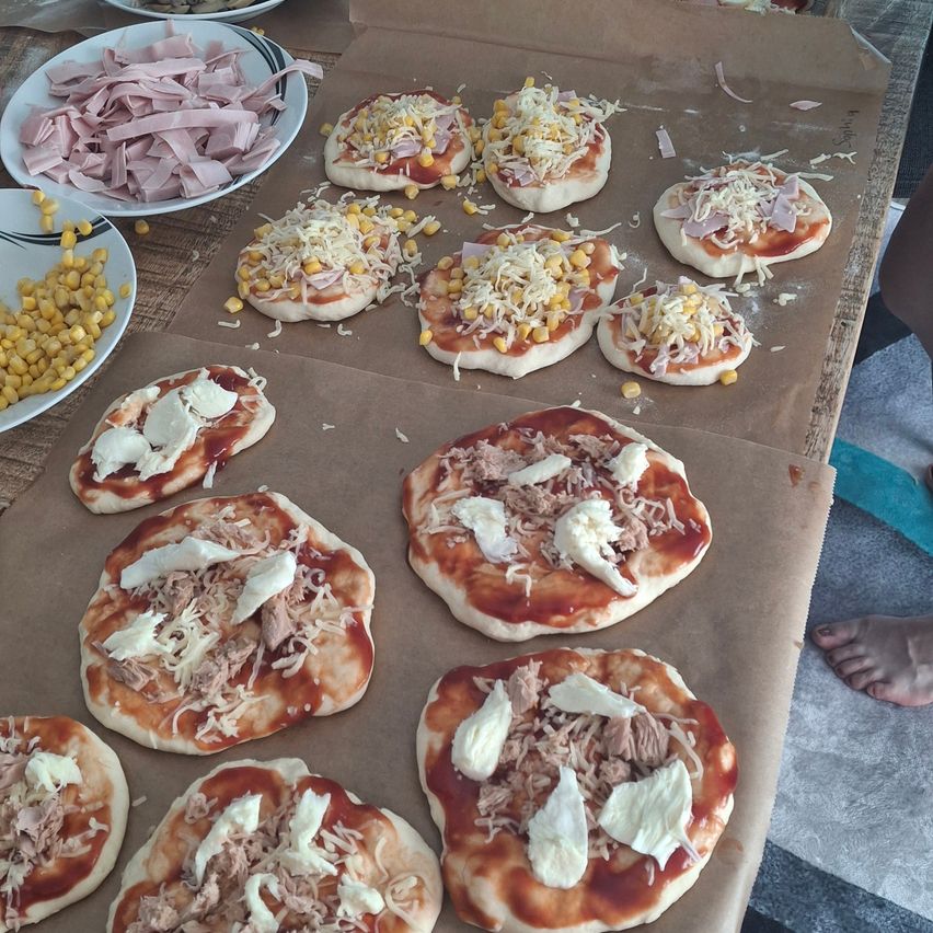 A table with various mini pizzas topped with cheese, corn, and ham, ready to be baked.