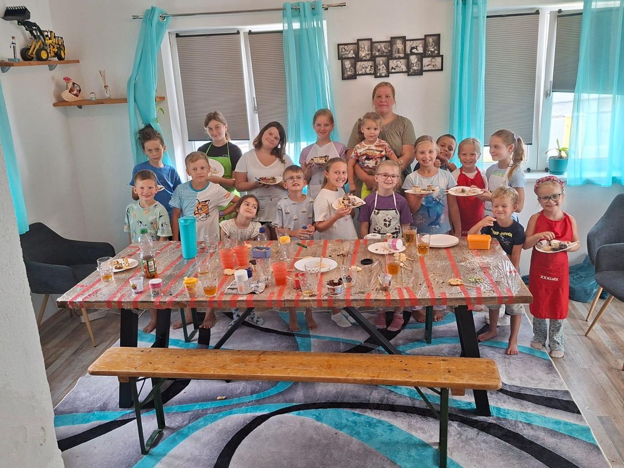 A group of children and adults gathered around a table, holding plates of food and drinks. They are smiling and posing for a photo. The table is covered with a plastic sheet and has various food items on it. The room has a carpet, a bench, and a wall with picture frames.