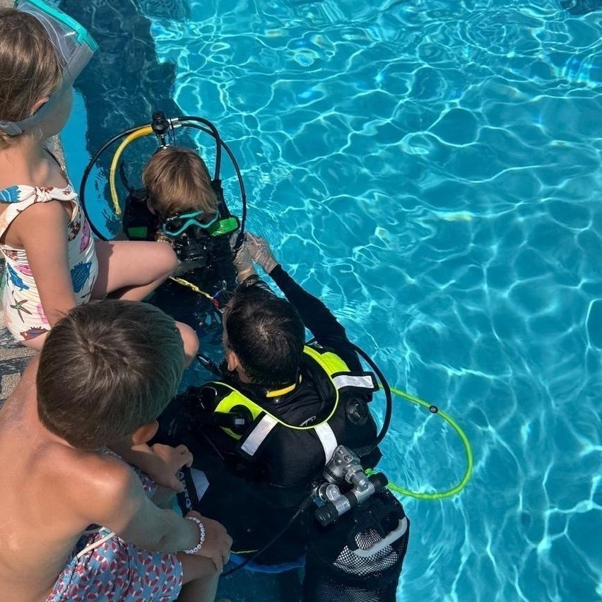 A scuba instructor is teaching a young child how to scuba dive in a pool with a few other children watching.