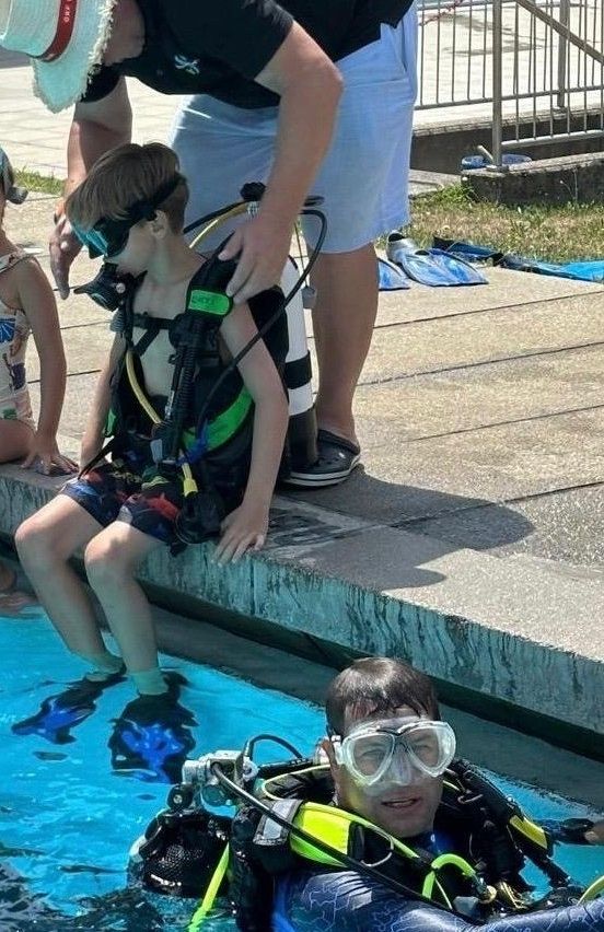 A young boy is getting ready to dive into a pool, wearing scuba gear and goggles. Another boy is already in the water, also in scuba gear. A person is standing on the pool's edge, holding the first boy's scuba equipment.