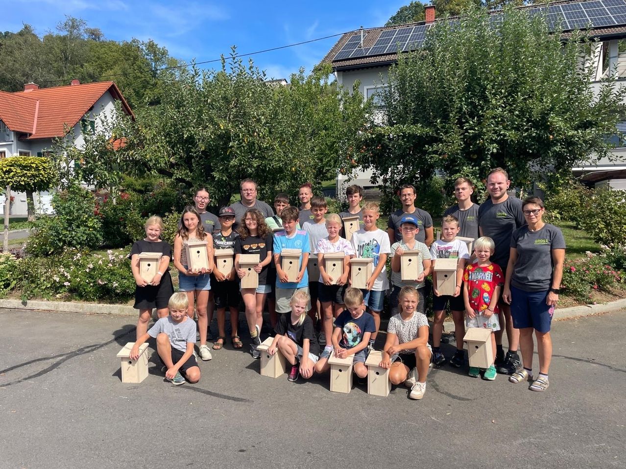 A group of children and adults pose for a photo with wooden birdhouses in a sunny outdoor setting.