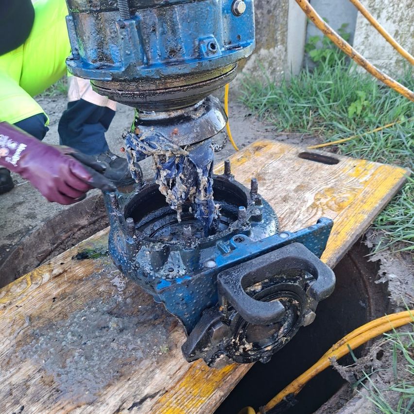 A person wearing gloves is working on a blue machine, possibly a pump, in a muddy area. The machine is placed on a wooden plank.