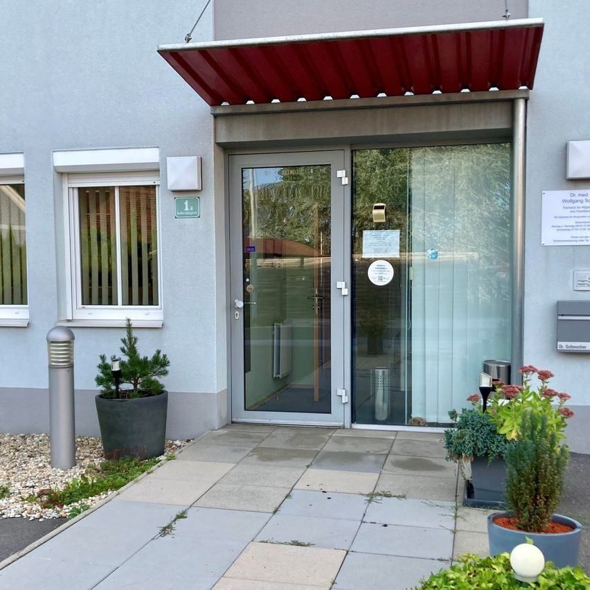 A modern building entrance with a glass door and red awning, featuring a small garden with potted plants and a mailbox on the right.