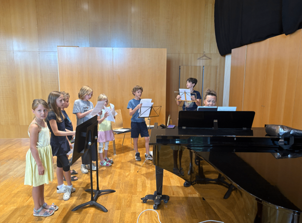 A group of children, dressed in casual attire, are standing in a room, holding music sheets and looking towards a piano. The room has wooden walls and floors.