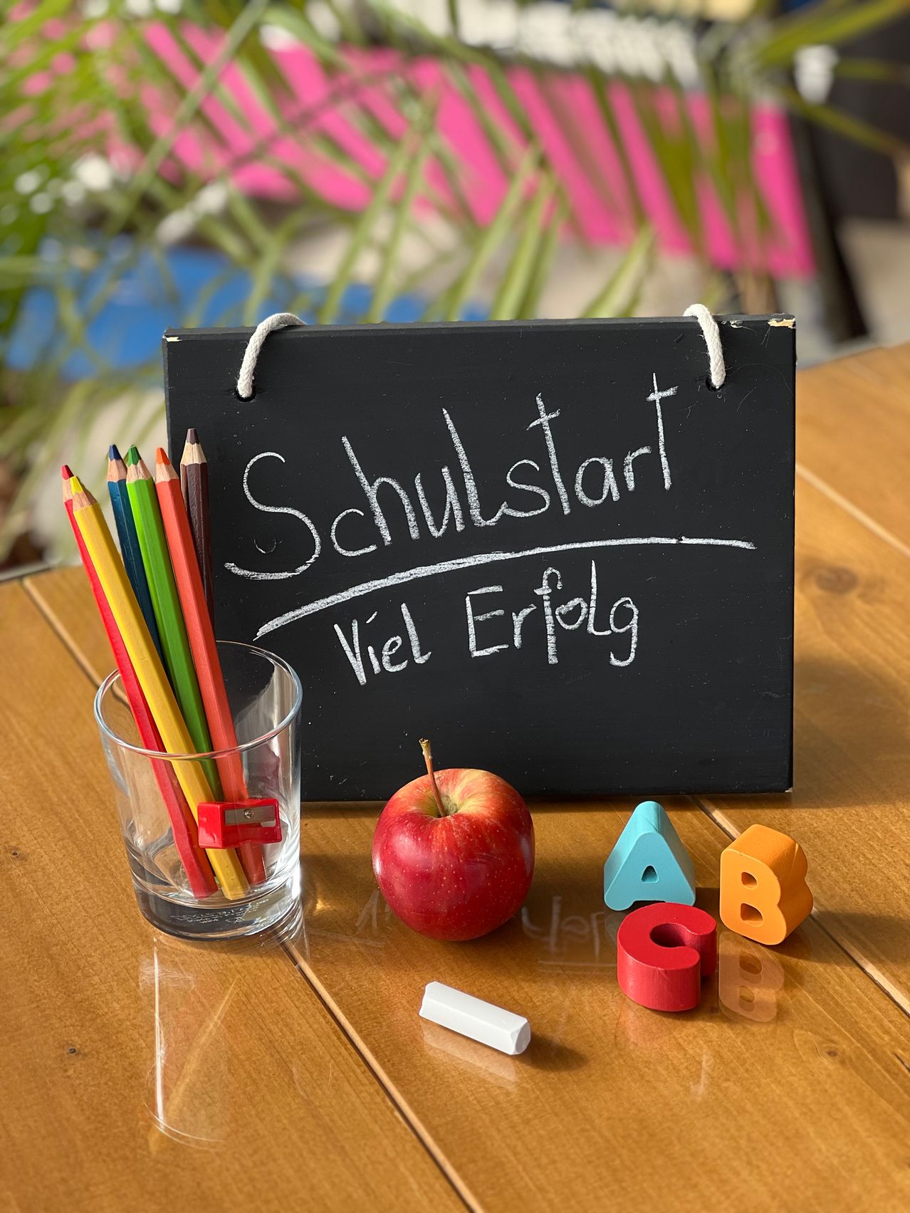 A blackboard with the words 'Schulstart Viel Erfolg' written on it, next to a glass of colored pencils, an apple, and alphabet blocks.