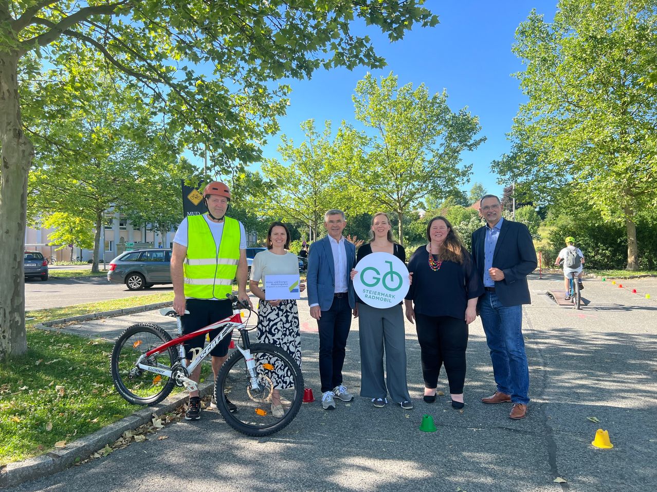 Group of people posing for a picture in front of a tree. One is holding a certificate and another holds a bike. All are smiling.