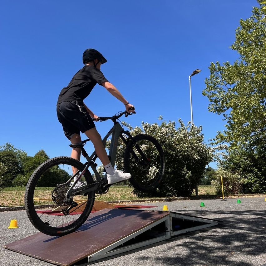 A person on a bike is performing a stunt on a ramp in an outdoor area. The person is wearing a helmet and sneakers. The area is surrounded by trees and has traffic cones.