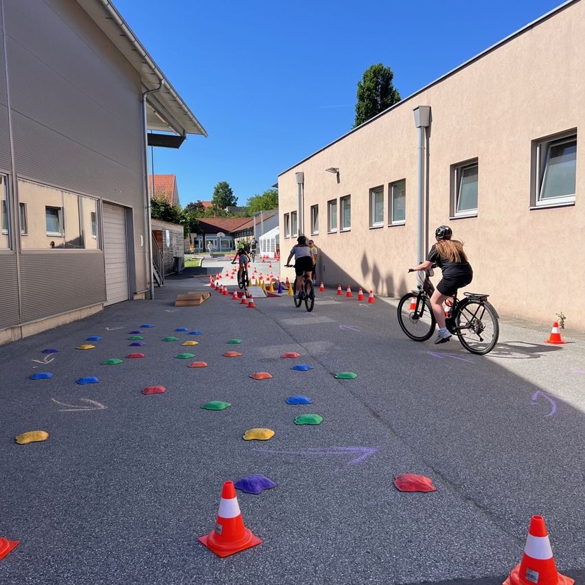 Three people ride bicycles through a course marked by colorful circles and orange cones. The course is set up between two buildings on a sunny day.