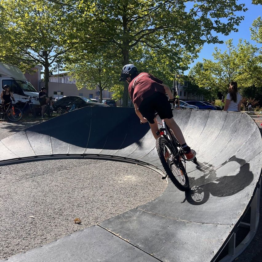 A boy rides his bike on a ramp. There are several people and vehicles around, including a parked white van. Trees and a building are in the background.