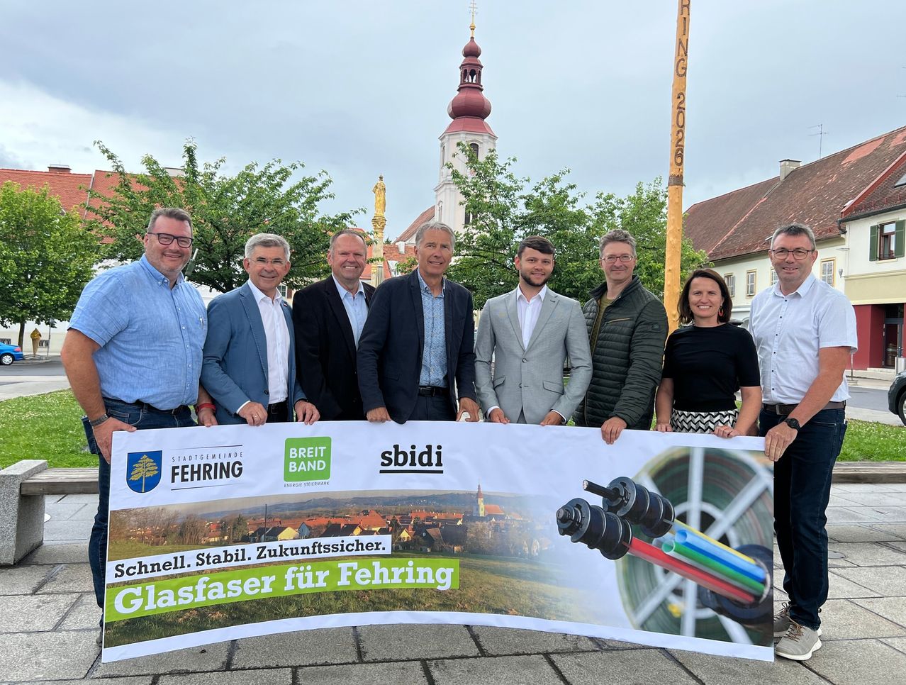 A group of men and women stand behind a banner with the text 'Schnell. Stabil. Zukunfstsicher. Glasfaser für Fehring'. They are standing in front of a building with a tower and trees.