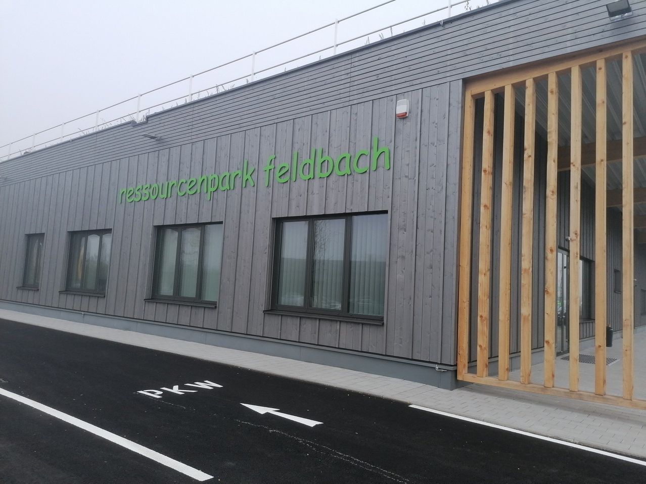 The front of a building with gray walls and windows, featuring the text 'ressourcenpark feldbach'. A wooden gate and a parking sign are visible.