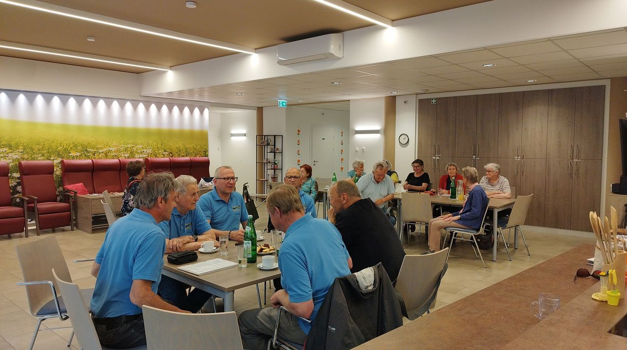 A group of people wearing blue shirts sit around a table in a room with white tiles, drinking and eating.