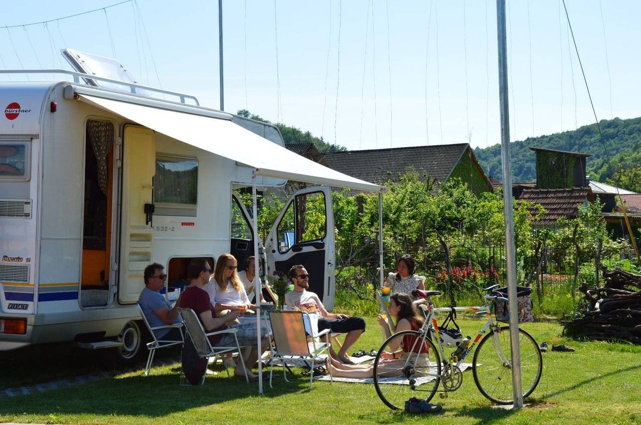 Eine Gruppe von Menschen sitzt um einen Tisch herum unter dem Vordach eines Wohnwagens. Einige haben Getränke und andere unterhalten sich. Ein Fahrrad und ein Haus sind im Hintergrund.