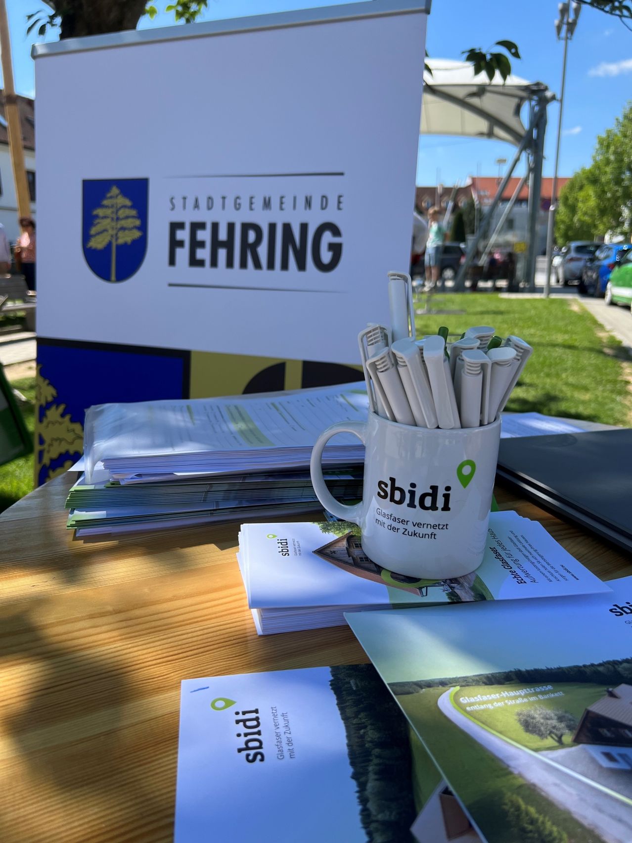 Ein Außentisch mit Werbematerialien der Stadtgemeinde Fehring. Ein Becher mit Stiften und eine Tasse mit der Aufschrift 'sbidi'. Broschüren und eine Karte liegen ebenfalls auf dem Tisch.