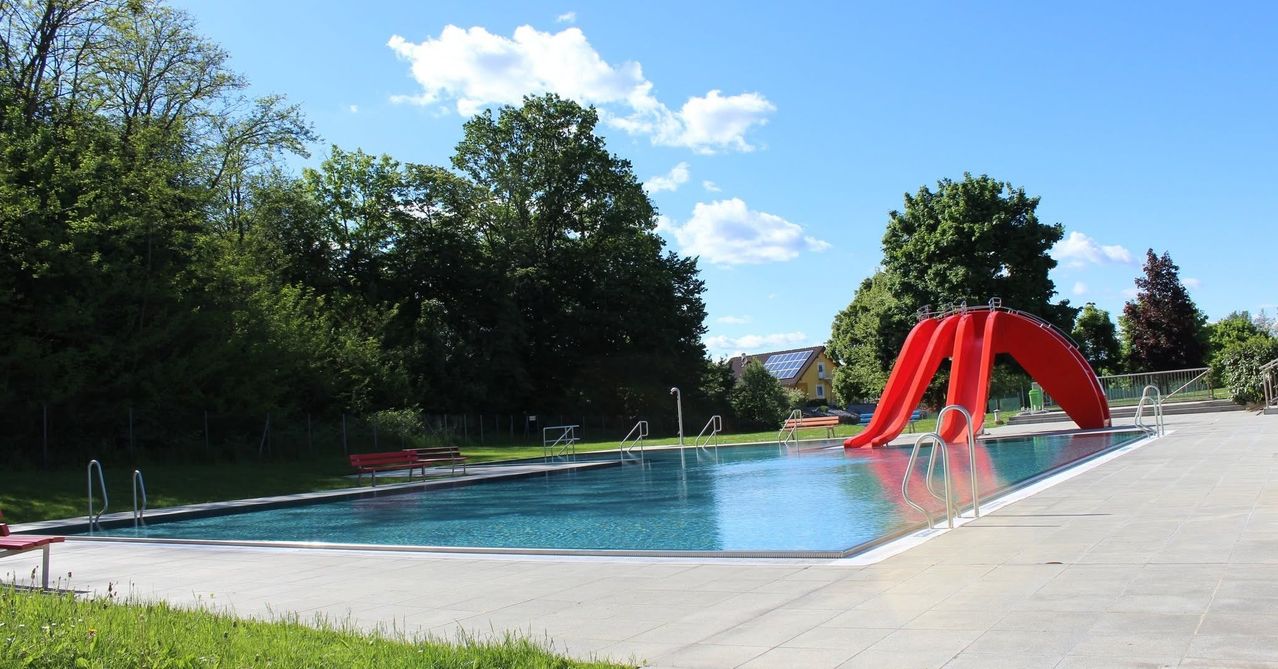 Ein Freibad mit einer roten Rutsche, umgeben von grünem Gras und Bäumen. Im Hintergrund ein Haus mit Solarzellen auf dem Dach.