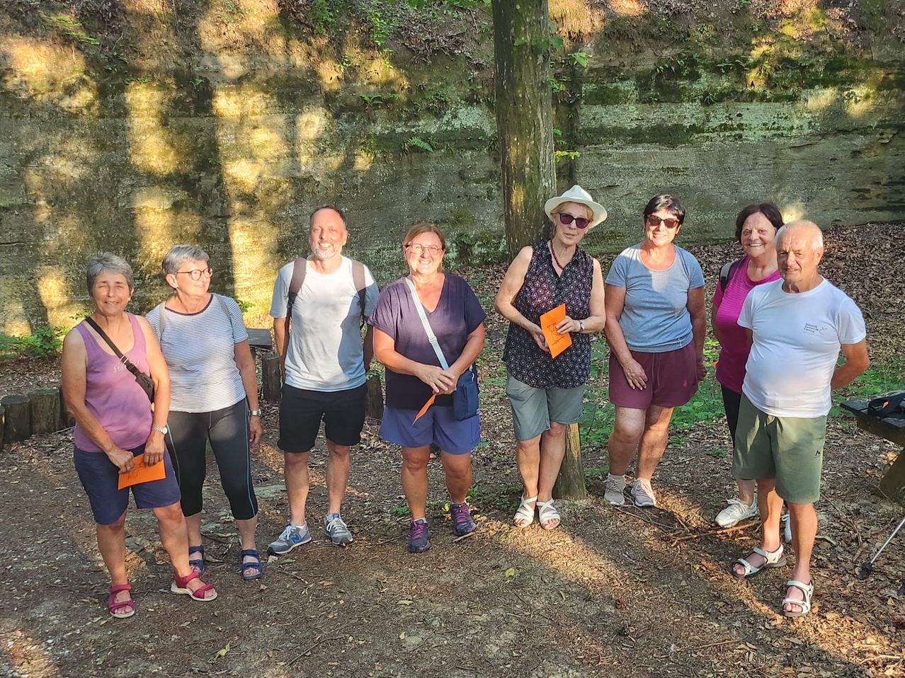 Eine Gruppe von Erwachsenen steht draußen und lächelt. Einige tragen Hüte und Brillen und halten Gegenstände. Sie befinden sich in der Nähe einer Steinmauer, mit Schatten von einem Baum.