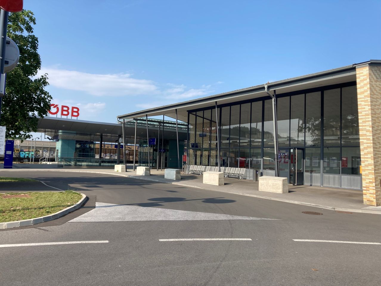 Eine leere Bahnsteigplattform mit einem blauen Schild und Bänken. Ein Gebäude mit Glaswänden und einem roten Schild, das 'OBB' steht.