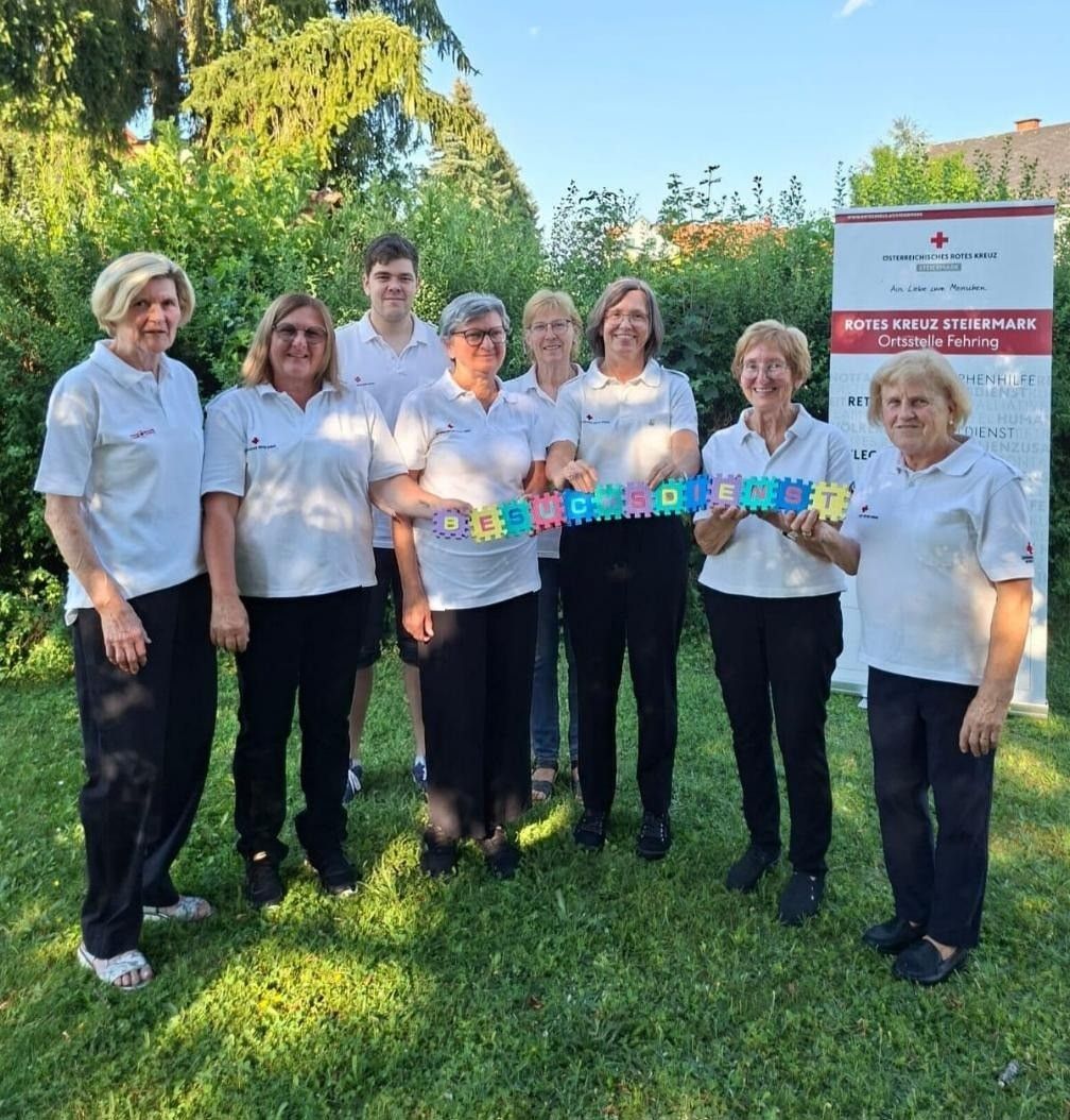 Eine Gruppe von Frauen und Männern in weißen Shirts und schwarzen Hosen steht auf einem Rasen und hält eine bunte Schleife. Dahinter steht ein Banner mit der Aufschrift 'Rotes Kreuz Steiermark Ortsstelle Fehring'.