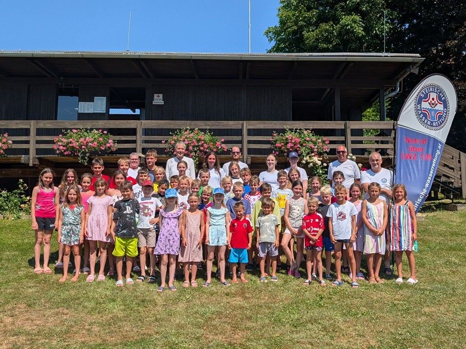 Eine Gruppe von Kindern und Erwachsenen posiert für ein Foto vor einem Gebäude mit Balkon und rosa Blumen.
